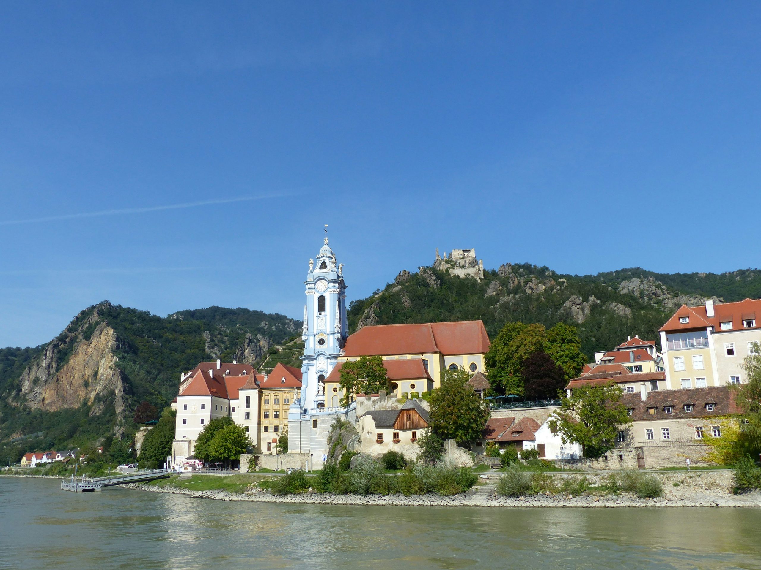 Blick auf Schloss Dürnstein von der Donau aus. Der Turm von Durnstein ist weiß blau und daher sehr auffällig. Das umgebende Gebäude ist gelb. Im Hintergrund sieht man bewaldete Hügel.