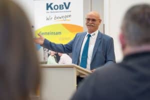 Ein fröhlich lachender Herr mit Glatze, Brille und Schnurrbart in blauem Anzug steht am Rednerpult vor dem KOBV-Rollup und gestikuliert weit mit der rechten Hand. Im Vordergrund sind die Hinterköpfe des Auditoriums.