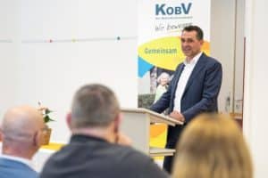 Ein dunkelhaariger Herr in schwarzem Anzug und weißem Hemd referiert an einem Rednerpult vor dem KOBV-RollUp. Im Vordergrund sind die Hinterköpfe des Auditoriums zu sehen.