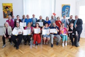 Eine Gruppe Menschen von etwa 20 Personen, vorne sitzend, hintere Reihe stehend, in einem sehr hellen, weißen Saal mit Fischgrätparkett.