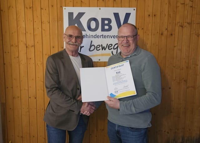 Zwei Männer mit Glatze und Brillen stehen vor einer Holzwand, auf der das KOBV Banner angebracht ist. Sie schütteln einander fröhlich lachend die Hände. Der Mann rechts im Bild hält sein Zertifikat in die Kamera.