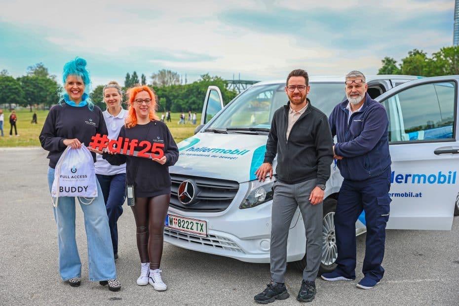 Eine Gruppe Menschen posiert vor einem KFZ der Marke Mercedes, im Hintergrund sind Bäume und Felder. Eine Dame mit roten Haaren hält ein rotes Objekt in Händen, das den Schriftzug #dif25 darstellt. Neben ihr steht eine Frau mit blauem Haar, die ein Stoffsackerl mit Logo der Firma FullAccess hält.