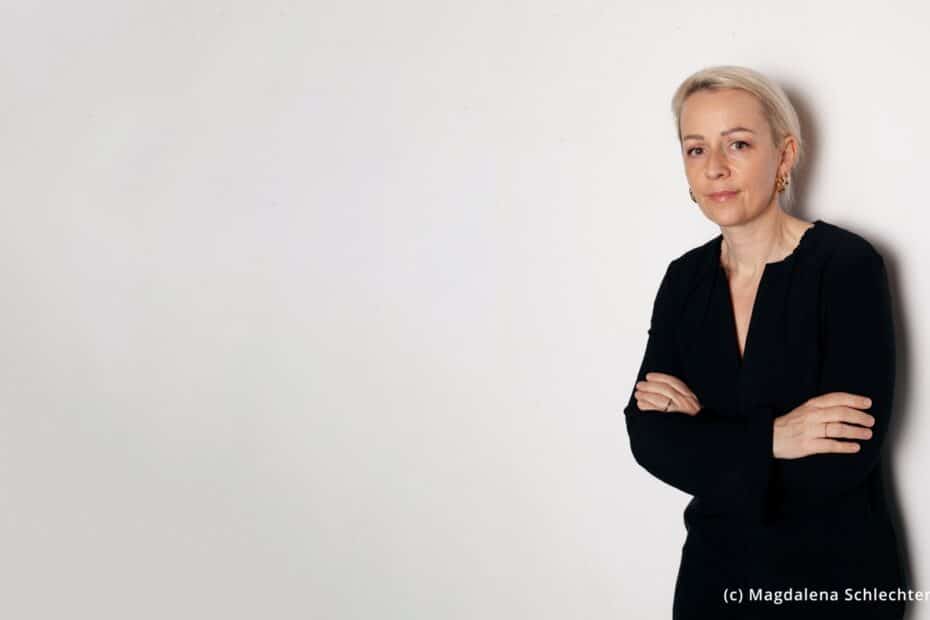 Eine blonde Frau in schwarzer Kleidung vor grauem Hintergrund.