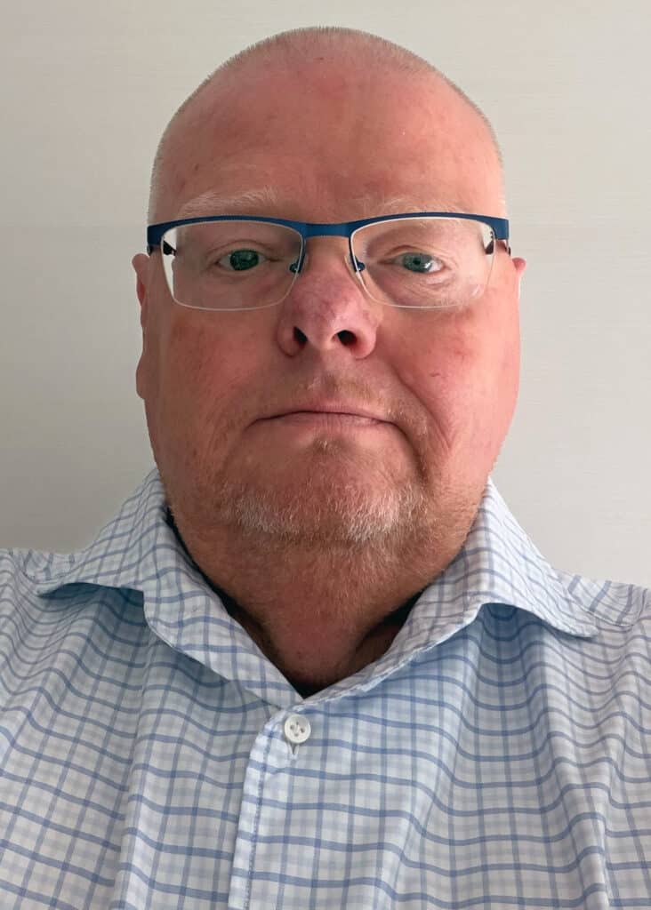 Porträtfoto eines Mannes mit sehr wenigen und sehr kurzen Haaren sowie einem kurzen Bart. Er trägt eine blaue, teils rahmenlose Brille.
